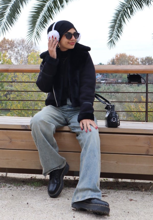Giacca nera corta con colletto in pelliccia, jeans blu spessi, scarpe nere chunky, cuffie bianche e soffici, e handbag nera accanto.