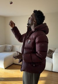 Homme en doudoune marron tenant un pot blanc, regardant vers le haut avec une main levée, debout dans un salon éclairé par la lumière du soleil.