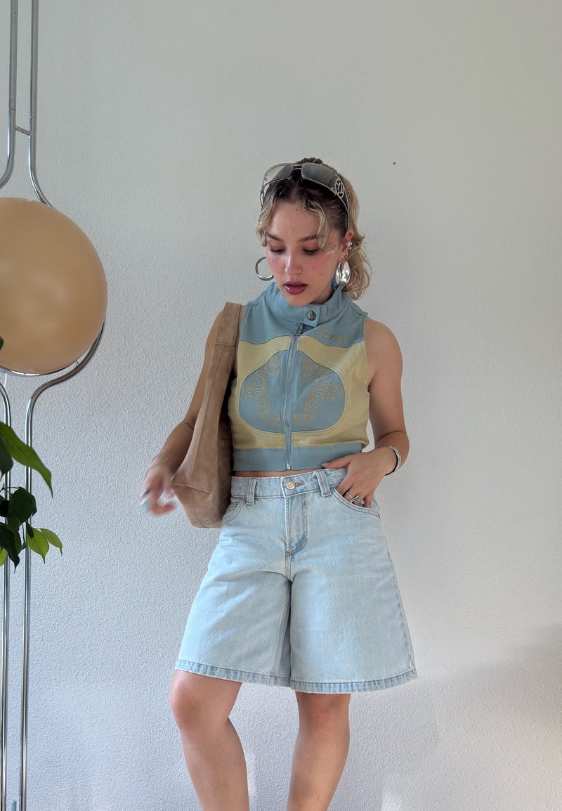 Jeune femme portant un top court sans manches et un short en denim clair, tenant un sac beige, debout à l'intérieur près d'une lampe et d'une plante.