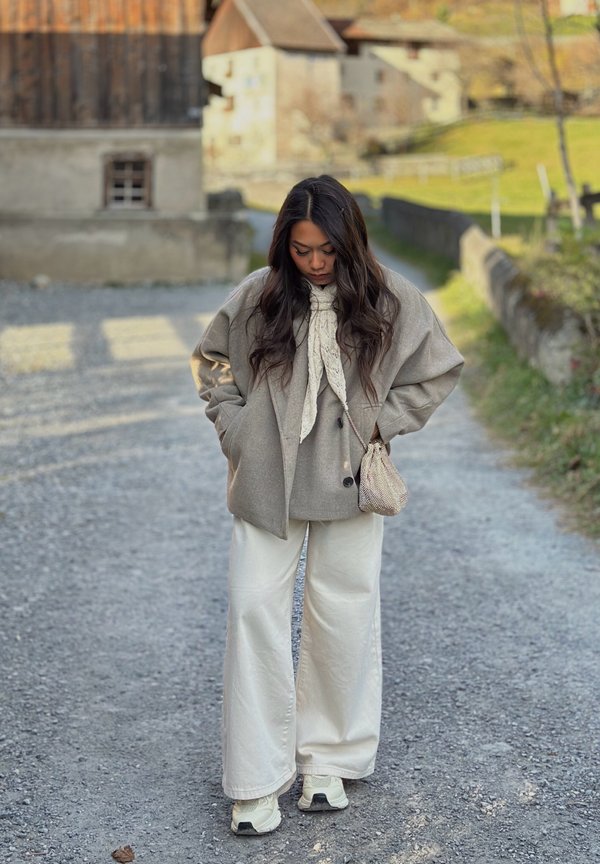 Oversize szara wełniana kurtka, kremowe spodnie o szerokich nogawkach, kremowy szal z wzorem, teksturowana torebka i białe trampki na żwirowej ścieżce w wiejskim otoczeniu.