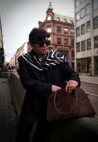 Smeđa torba od brušene kože s zaobljenim vrhom i dvostrukim ručkama. Osoba nosi crno-bijelu prugastu šal i crnu jaknu, stojeći na gradskom ulici.