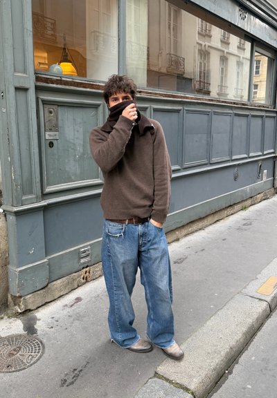 Hombre de pie en la acera junto a un edificio gris, lleva un suéter marrón cubriéndose la boca y vaqueros azules holgados con zapatos desgastados.