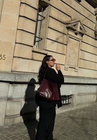 Sac à épaule bordeaux avec une finition texturée, porté par une personne en tenue noire, se tenant contre un mur en pierre. La lumière du soleil projette une ombre.