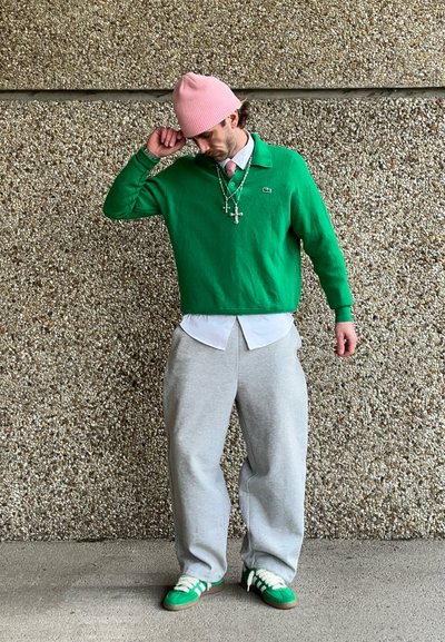 Suéter verde sobre una camisa blanca, pantalones de chándal grises y zapatillas verdes con acentos blancos. Un gorro rosa y collares en capas completan el look.