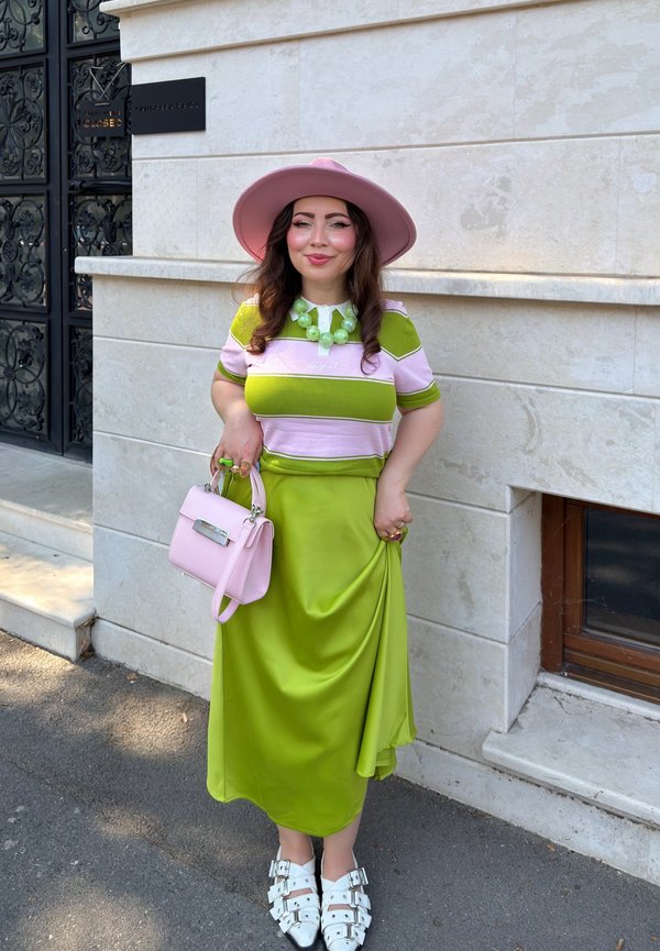 Femme portant un chapeau rose à large bord, une chemise rayée verte et rose, une jupe vert citron, des chaussures blanches à boucle, tenant un sac à main rose, se tient près d’un mur en pierre.