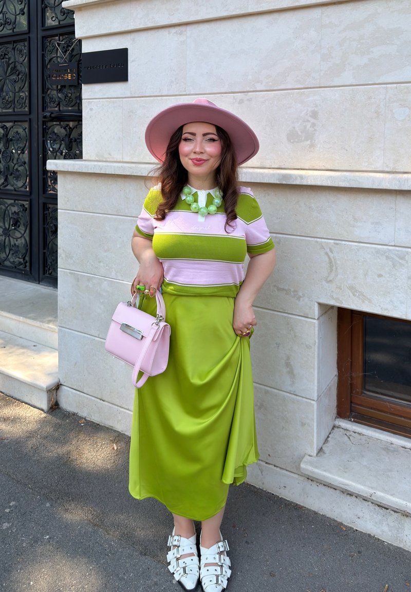 Woman in a pink wide-brim hat, green and pink striped shirt, lime green skirt, white buckled shoes, and holding a pink handbag stands by a stone wall.