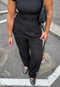 Persona che indossa pantaloni neri, una maglietta nera attillata, ballerine nere con borchie e porta una piccola borsa nera, in piedi sull'asfalto vicino a una ringhiera metallica.