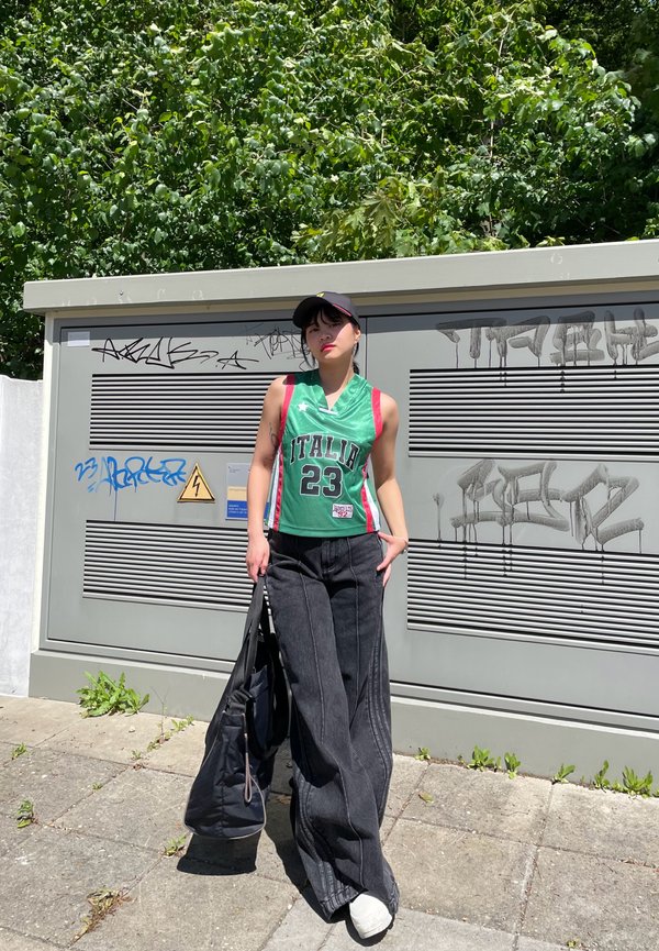 Groene basketbaljersey met "ITALIA" en "23," gecombineerd met brede zwarte denim broek en een zwarte pet, tegen een achtergrond van graffiti.
