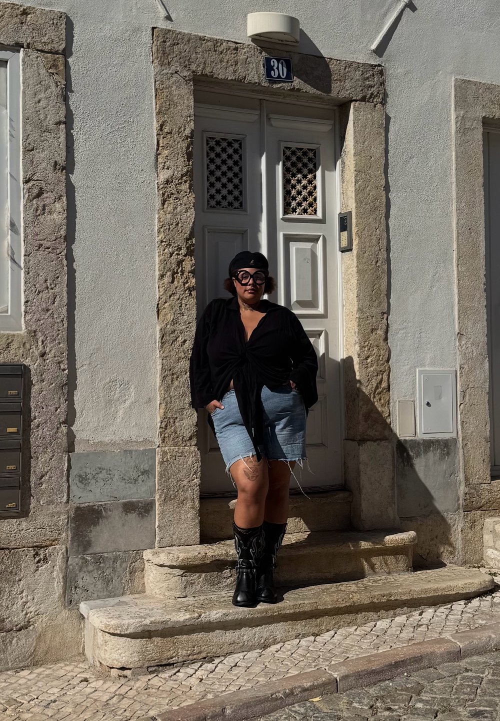 Chemise noire nouée devant, short en jean déchiré, et bottes de cowboy noires avec des accents de flammes, debout près d'un mur de pierre texturé et d'une porte blanche.