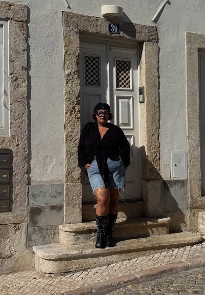 Camisa negra anudada al frente, pantalones cortos de mezclilla desgastados y botas vaqueras negras con detalles de llamas, junto a una pared de piedra texturizada y una puerta blanca.