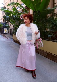 Femme aux cheveux bouclés portant une veste blanche duveteuse, un crop top gris, une jupe en satin rose et des bottes marron, tenant un sac à main à motifs et un téléphone.