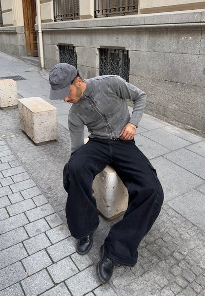 Sudadera gris con cierre, con un patrón texturizado, pantalones cargo negros de gran tamaño y botas negras, sentado en un banco de piedra en un entorno urbano.