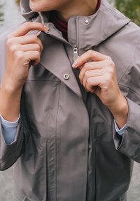 Grå vattentät jacka med hög krage, med dragkedja fram och tryckknappar, händer som justerar dragkedjan medan de är utomhus.