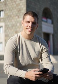 Světle béžové žebrované tričko s dlouhým rukávem a vyšitým logem, sedící postava s chytrým telefonem, tmavé džíny, rozmazané kamenné pozadí.