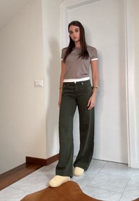 Grijze t-shirt met korte mouwen en witte bies, olijfgroene wijde broek en beige slippers, gepositioneerd tegen een eenvoudige muur.