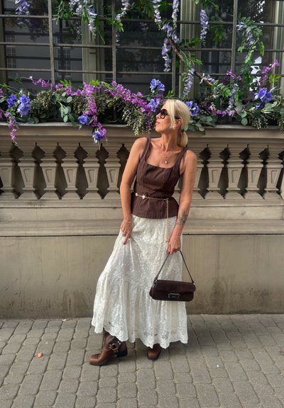 Top marrón ajustado con tirantes y una cadena, falda maxi blanca de encaje, botines marrones, y un bolso oscuro. Fondo floral con vegetación.