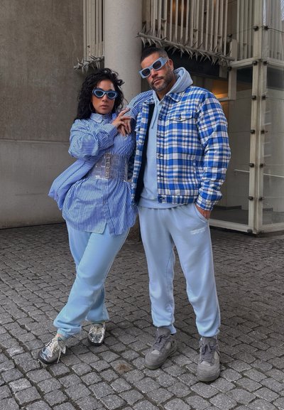 Camisa de rayas azul y blanca de gran tamaño, joggers azul claro y zapatillas grises. Los accesorios incluyen gafas de sol azules y una chaqueta a cuadros.