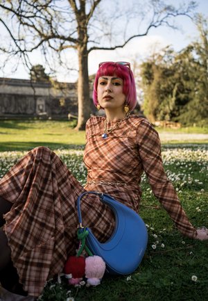 Joven mujer con cabello rosa que lleva un vestido marrón a cuadros, sentada en el césped con un bolso azul y flores de margarita en un entorno de parque.