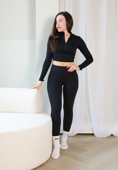 Top corto atlético negro con collar de cremallera, combinado con leggings negros de cintura alta. Sofá en forma de tazón blanco texturizado en el fondo.