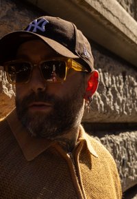 Homme barbu portant une casquette noire NY, des lunettes de soleil ambrées reflétant une scène de rue, des boucles d'oreilles en forme de cercle et de croix, et un pull zippé marron.