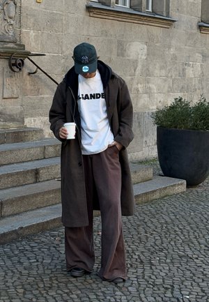 Abrigo largo marrón, camiseta blanca con letras oscuras, pantalones marrones sueltos, zapatos negros, gorra verde. Sosteniendo una taza de café, de pie en unos escalones de piedra.