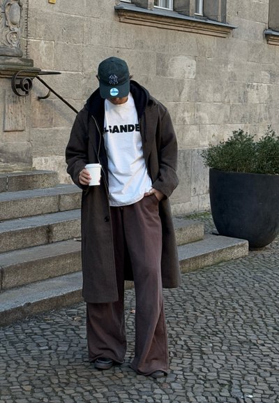 Abrigo largo marrón, camiseta blanca con letras oscuras, pantalones marrones sueltos, zapatos negros, gorra verde. Sosteniendo una taza de café, de pie en unos escalones de piedra.
