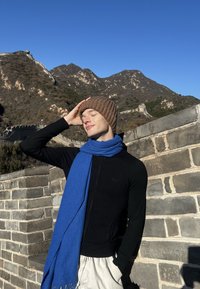 Črni pulover z zadrgo in modro ruto, ki ju nosi oseba z rjavo pleteno kapo, postavljena ob kamniti steni z goratim ozadjem.