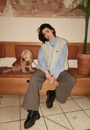 Chaleco sin mangas de color gris claro sobre una camiseta azul, con pantalones anchos marrones y botas negras. Un perro de color beige está sentado al lado de la persona en un banco de madera.