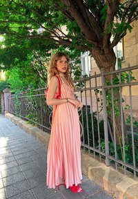 Maxikleid mit Faltenwurf in einem Farbverlauf von Pink, aus leichtem Stoff gefertigt. Kombiniert mit roten Sandalen und passenden Ohrringen, umgeben von viel Grün.