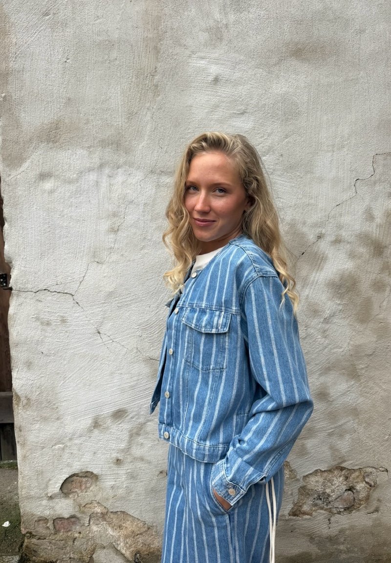 Femme blonde en veste et jupe rayées bleu et blanc, debout de profil, une main dans la poche, contre un mur beige usé.