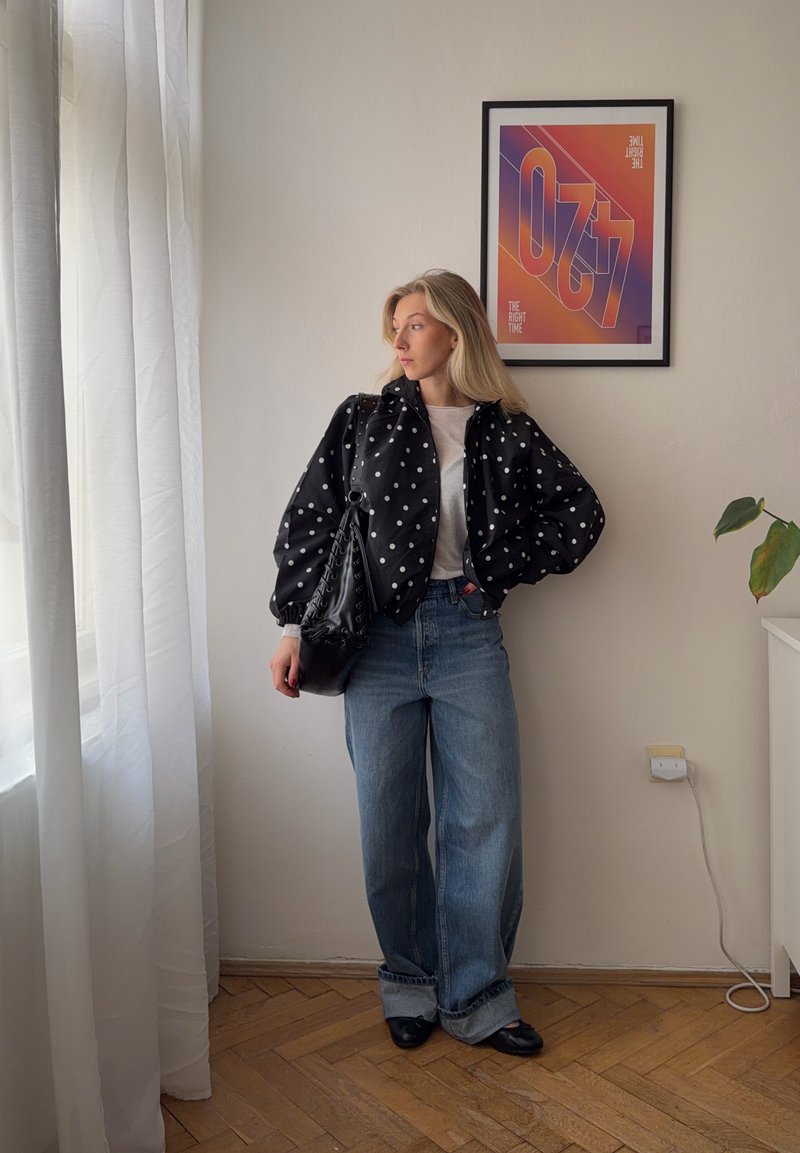 Femme aux cheveux blonds portant une veste noire à pois, une chemise blanche, un jean large bleu, des chaussures noires, tenant un sac noir, debout à l'intérieur près d'une fenêtre.