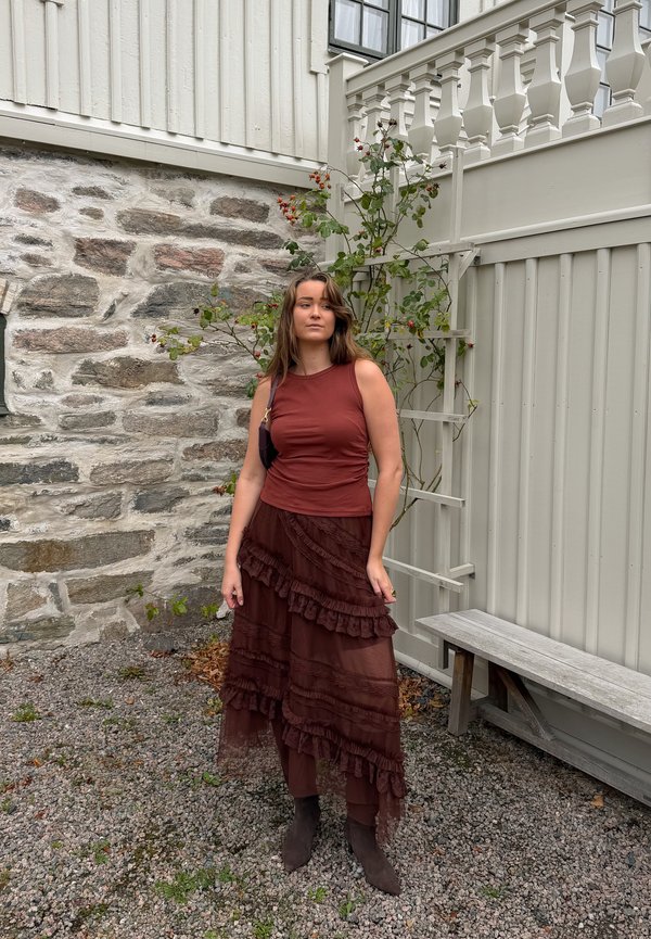 Una mujer con una blusa de color rojo óxido y una falda marrón de varias capas está de pie en un patio de grava, junto a un muro de piedra, una planta trepadora en flor y un banco de madera.