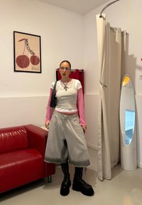 Hallid üleulatuvad lühikesed püksid nööriga, roosad pika varrukaga pluusid halli vestiga, millele on lisatud mahukad mustad saapad, seistes minimalistlikus toas.