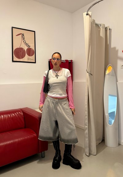 Pantalones cortos grises extragrandes con cordón, mangas largas rosas debajo de una camiseta sin mangas gris, combinados con botas negras gruesas, de pie en una habitación minimalista.