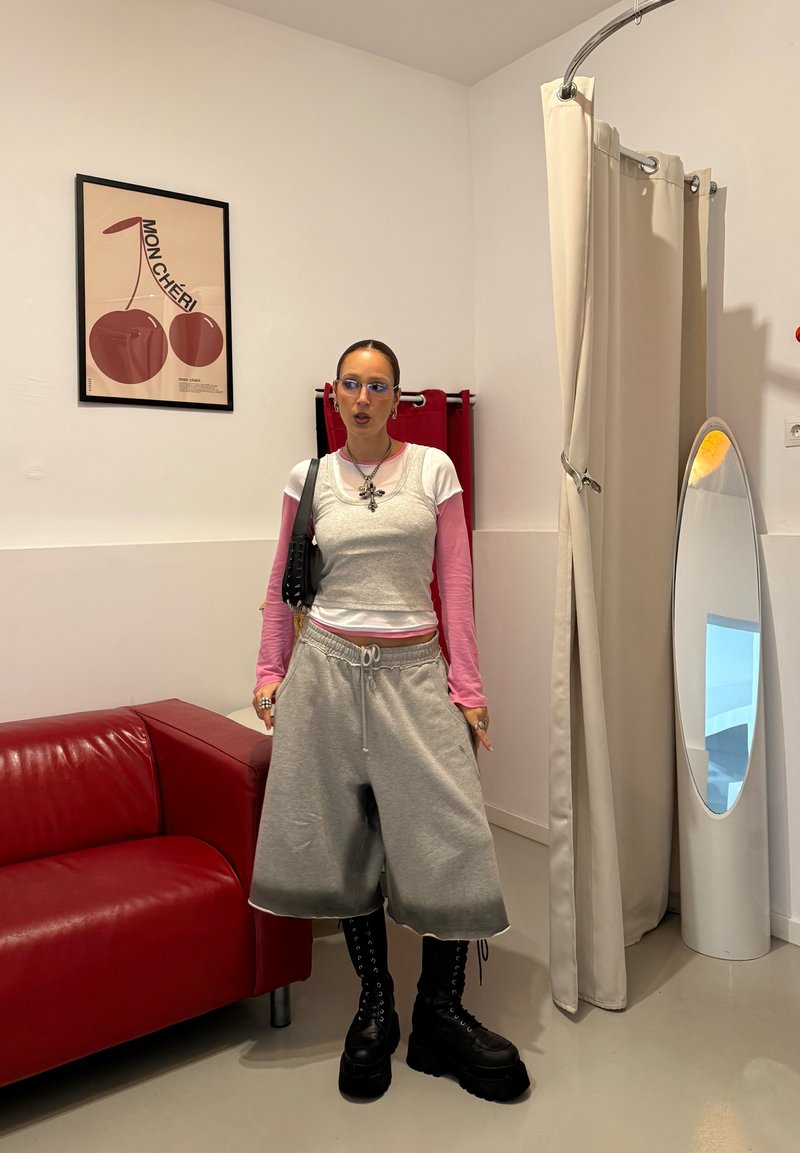 Shorts gris oversized avec cordon de serrage, manches longues roses sous un débardeur gris, assortis à des bottes noires épaisses, debout dans une pièce minimaliste.