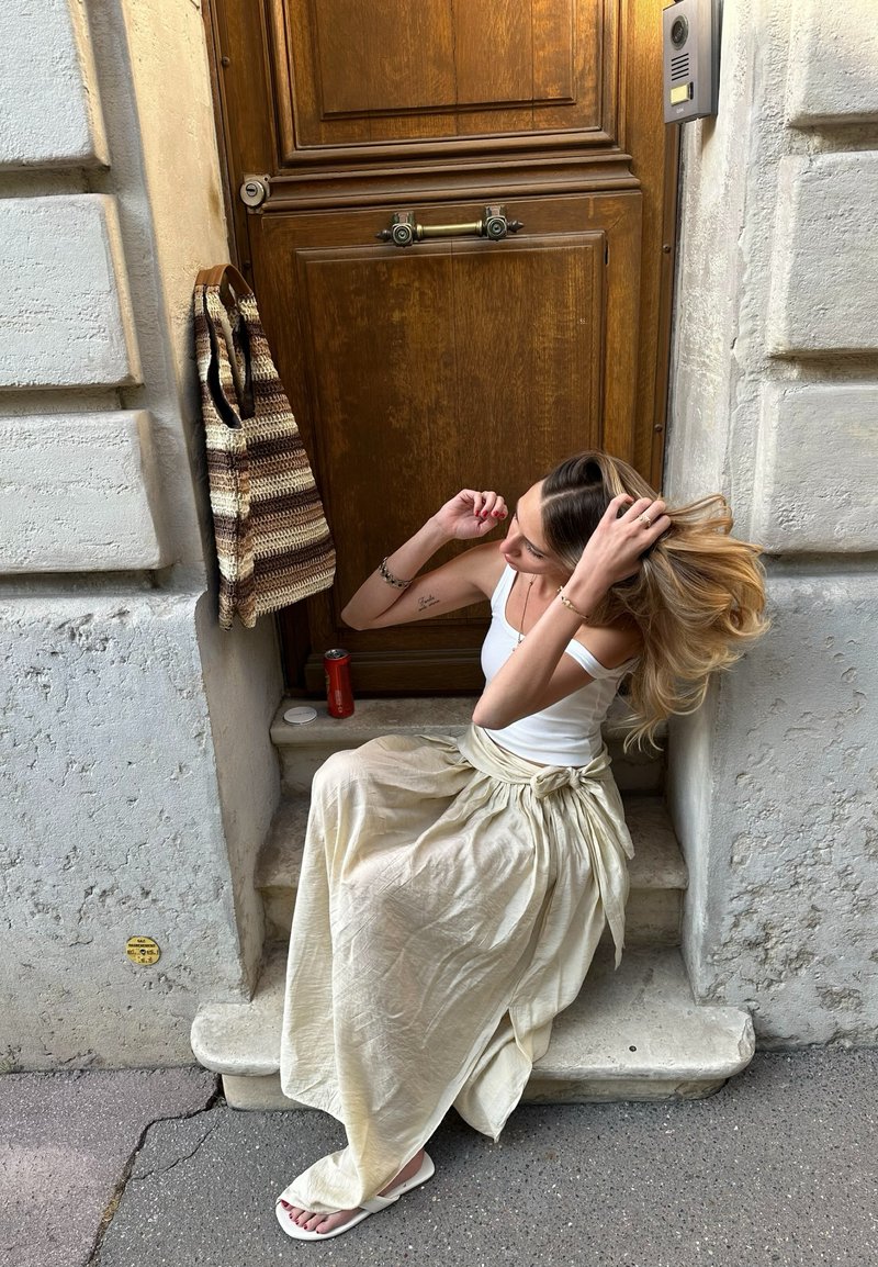 Femme en débardeur blanc et longue jupe beige assise sur le seuil en pierre, arrangeant ses cheveux, à côté d'un sac rayé accroché au mur et d'une porte en bois.