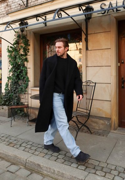 Abrigo negro, suéter negro, jeans de un azul claro y zapatos negros que lleva un hombre mientras camina por un camino de piedra fuera de un café rodeado de vegetación.