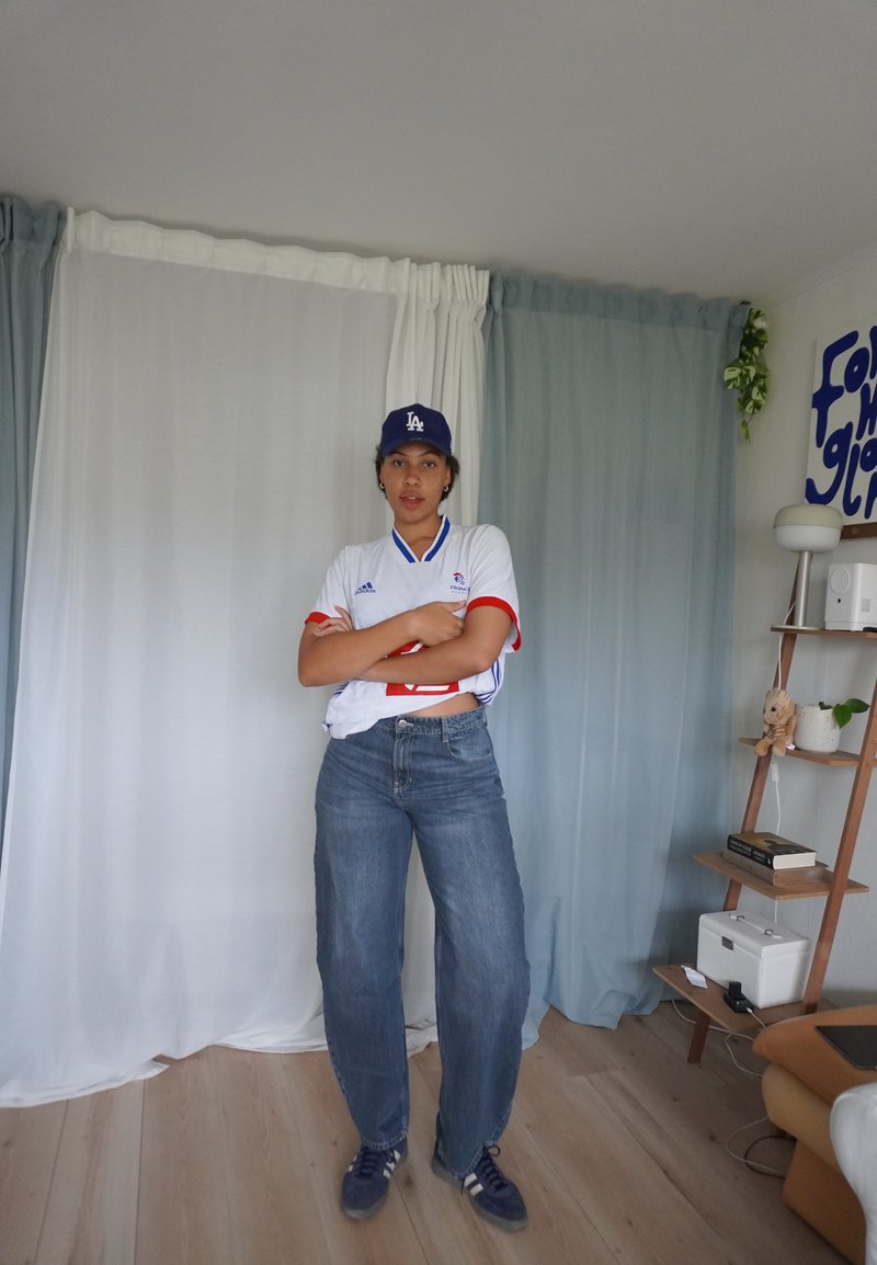 Jersey bianco a maniche corte con accenti rossi e blu, abbinato a jeans blu larghi e un cappellino blu scuro. In piedi su un pavimento in legno chiaro.