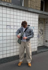 Grauer Zip-Hoodie, weißes T-Shirt, beigefarbene weit geschnittene Hose und gelbe Stiefel. Trägt eine lässige grüne Tasche. Texturierten Fliesen Hintergrund.