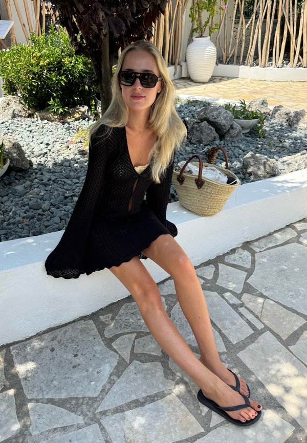 Frau mit langen blonden Haaren, Sonnenbrille, schwarzem Kleid und Flip-Flops sitzt auf einem weißen Steinsims nahe einem felsigen Garten mit einer Strohtasche.