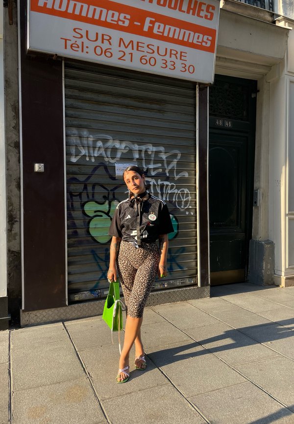 Svart skjorte med grafikk, leopardmønstrede capribukser, grønn håndveske og sandaler med rosa stropper. Urban bakgrunn med graffiti.