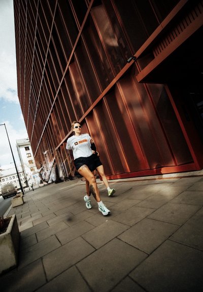 Dos personas corriendo junto a una acera de la ciudad al lado de un edificio moderno alto de color cobre en un día soleado.