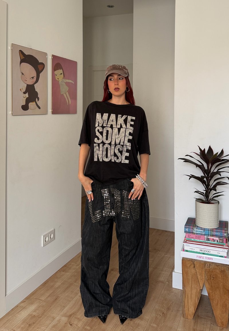 Young woman wearing a "MAKE SOME NOISE" shirt, studded pants, and a cap, standing in a room with art on walls and a plant on a table.