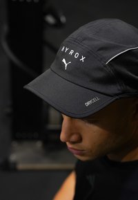 Νεαρός άνδρας που φοράει μαύρο καπέλο Puma HYROX με ύφασμα DRYCELL, κοιτάζοντας προς τα κάτω σε εσωτερικό χώρο με χαμηλό φωτισμό.