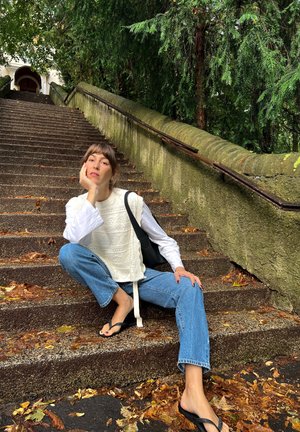 Top de punto blanco texturizado superpuesto a una camiseta de manga larga, combinado con jeans de mezclilla azules y chanclas negras, sentado en escalones de piedra.