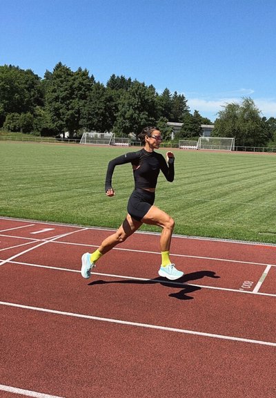 Atleta corriendo en una pista roja, vistiendo una camiseta de manga larga negra, pantalones cortos negros, calcetas amarillas brillantes y zapatillas de running azul claro.
