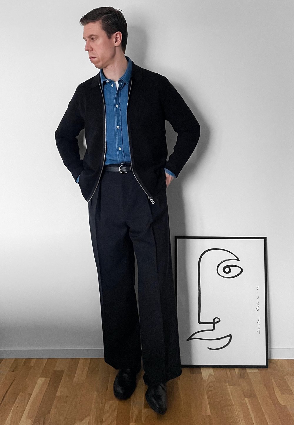 Homme en veste noire et pantalon large noir se tient près d'une œuvre d'art minimaliste encadrée représentant un visage en lignes, posée sur un sol en bois contre un mur blanc.