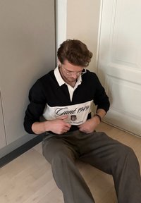 Chemise de style rugby pour hommes, noire et blanche, avec le texte "Gant 1949", manches longues et col contrastant. Assis sur un sol en bois dans une pose décontractée.