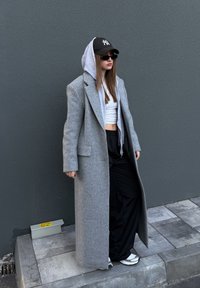 Manteau en laine gris oversize sur un hoodie gris, un crop top blanc et un pantalon noir large. Une casquette noire et des lunettes de soleil complètent le look.