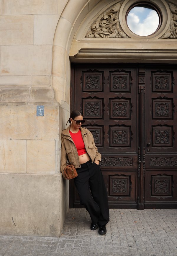 Mujer con gafas de sol, top corto rojo, chaqueta marrón y pantalones negros apoyada contra una pared de piedra cerca de una puerta de madera ornamentada con detalles tallados.